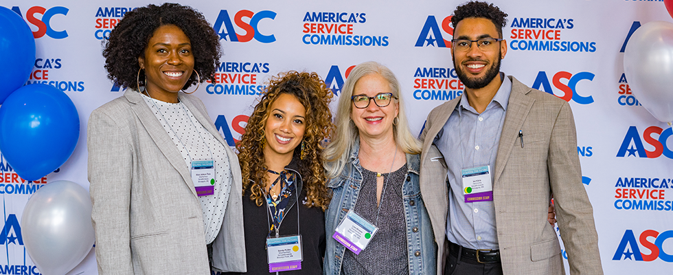 Decorative image. Four people pose for a photo in front of an ASC step-and-repeat background. They are all looking at the camera and smilling.