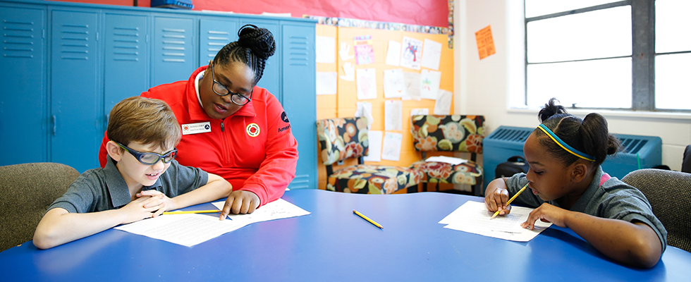 Decorative image. An AmeriCorps member helps two students with their assignment at a table.